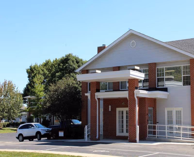Front entrance of a two-story brick senior living building with columns, an accessibility ramp, and parked cars under a clear sky.