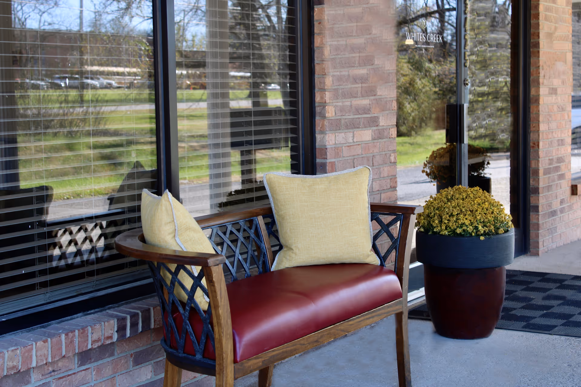 A bench with yellow pillows and a potted plant sits outside the glass entrance of Whites Creek Wellness & Rehabilitation Center.
