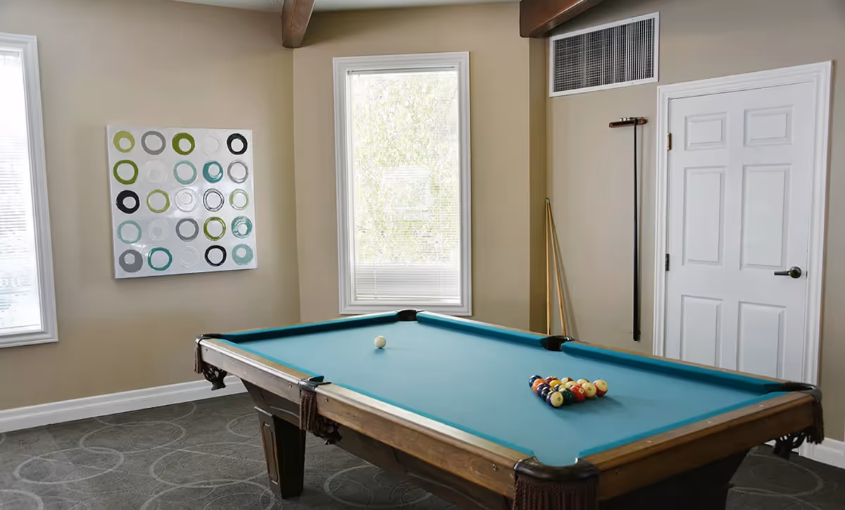 A well-lit interior recreation room featuring a pool table with racked balls, cue sticks, and wall art between two windows.