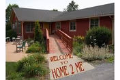 Single-story red building with a ramp leading to the entrance, surrounded by greenery and outdoor seating. A sign in front reads 'WELCOME TO HOME 2 ME'.