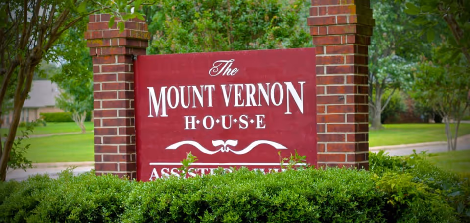 A red sign with white text reading 'The Mount Vernon House' mounted between two brick pillars, surrounded by green bushes and trees in a landscaped outdoor setting.