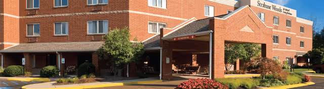 Exterior view of Strabane Woods of Washington senior living facility showing a multi-story brick building with several windows, a covered entrance with seating, landscaped bushes, trees, and a clear blue sky.