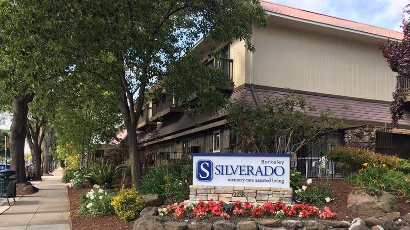 Exterior view of Silverado Berkeley Memory Care Community building with a sign in front surrounded by flowers and landscaping. Trees line the sidewalk next to the building under a partly cloudy sky.