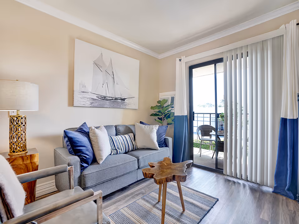 Bright living room with a gray sofa, nautical wall art, wooden coffee table, and sliding glass door opening to a small balcony.