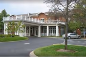 Front entrance of a brick senior living building with a covered porte-cochère, balcony railing, circular driveway, and parked cars.