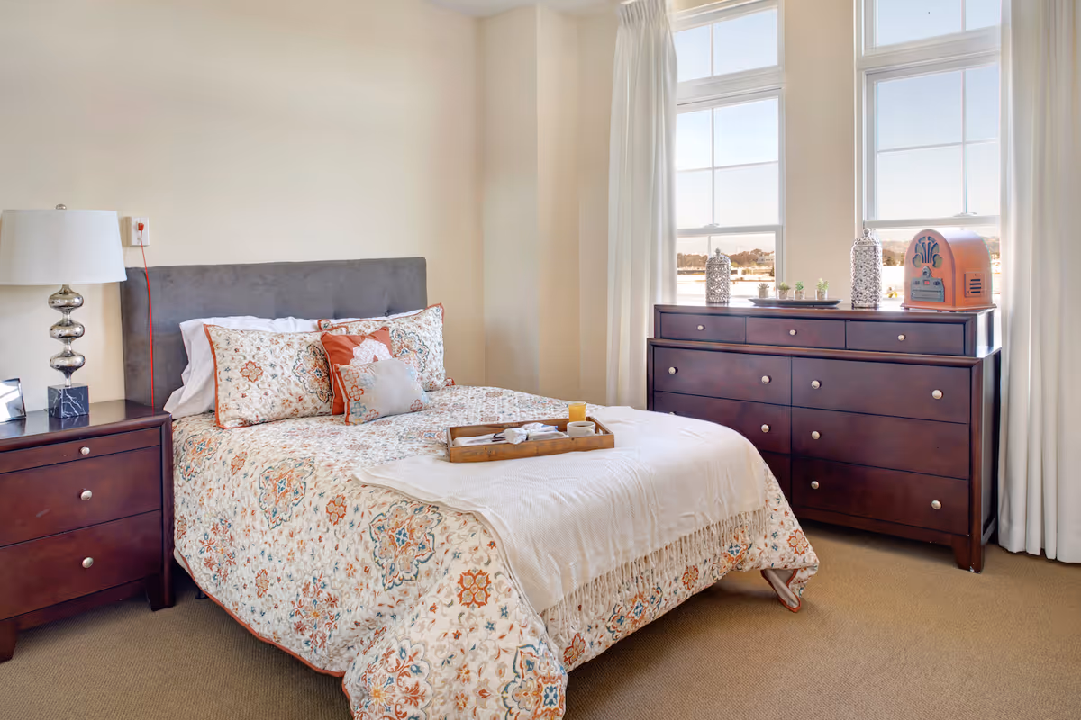 A cozy bedroom with a bed featuring a patterned quilt and multiple pillows. There is a wooden nightstand with a modern lamp on the left side of the bed. On the right side, a large wooden dresser is positioned under two tall windows with white curtains, decorated with small plants and decorative items.