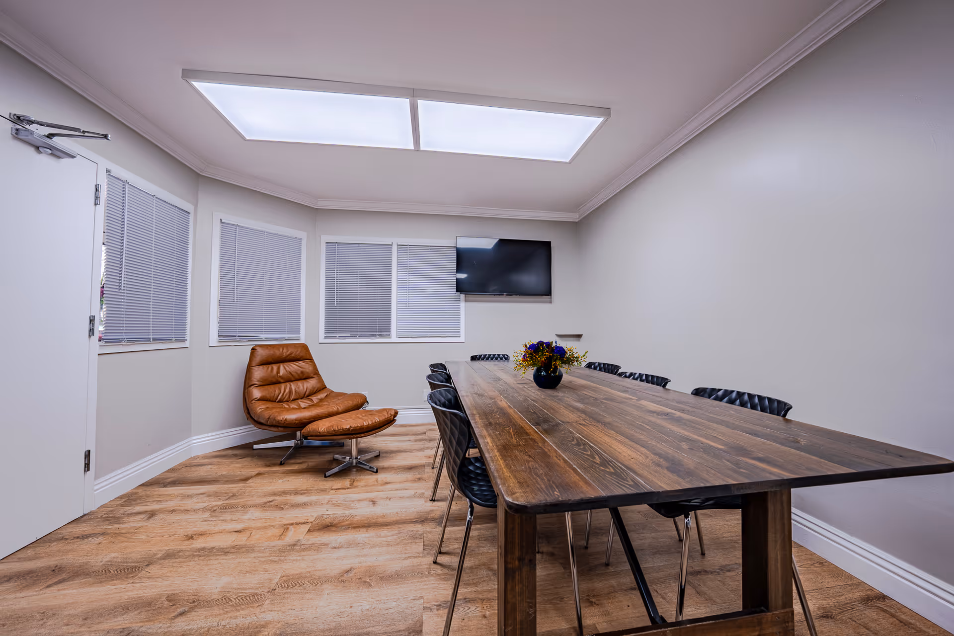 A modern meeting or dining room with a long wooden table surrounded by black chairs. A brown leather lounge chair with an ottoman is positioned near three windows with closed blinds. A flat-screen TV is mounted on the wall above the table. The room has light-colored walls, wood flooring, and a large rectangular ceiling light.