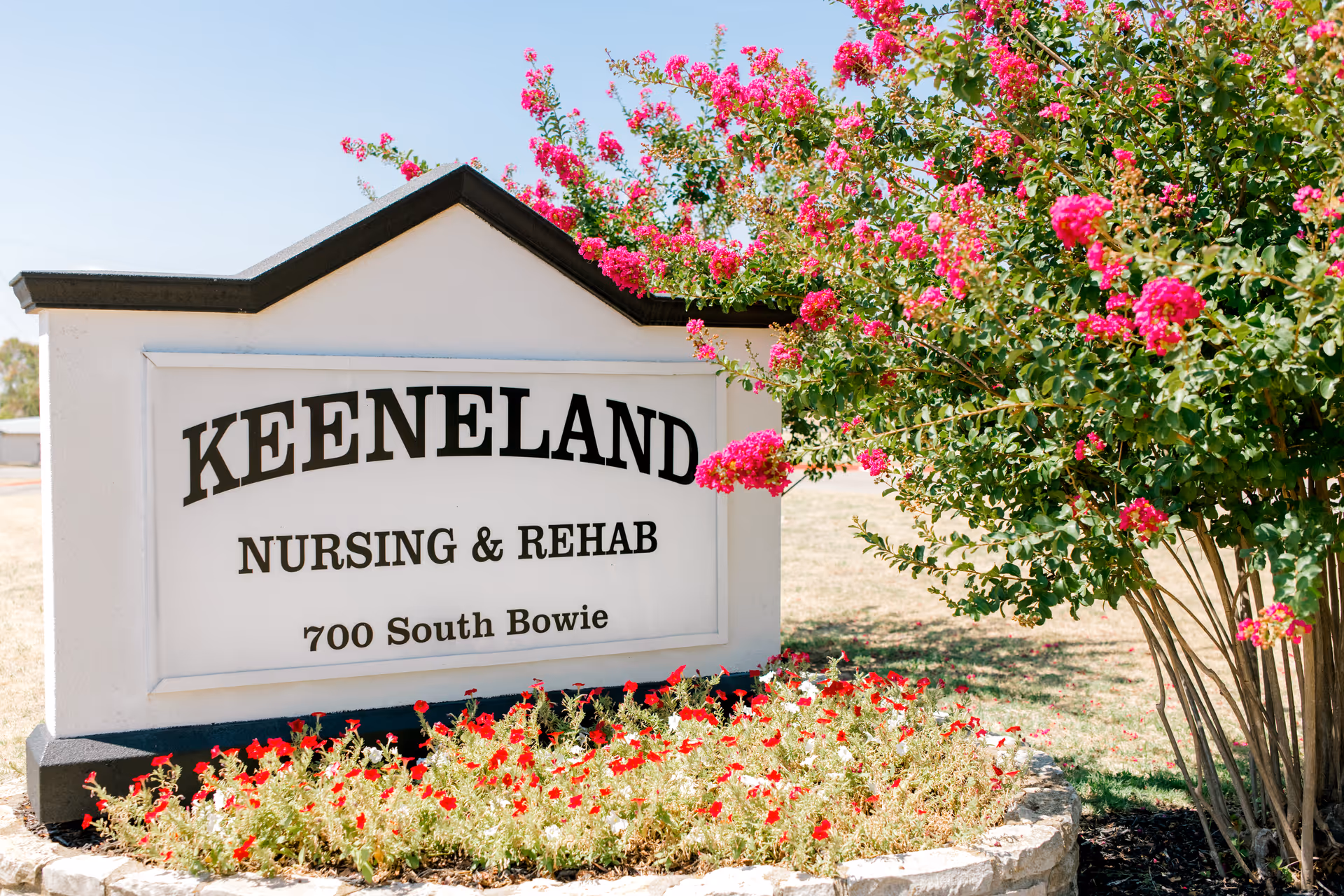 Outdoor sign for Keeneland Nursing & Rehab located at 700 South Bowie, surrounded by blooming pink flowers and greenery under a clear sky.