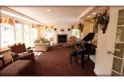 A cozy living room with patterned carpet, a grand piano, two armchairs, a sofa, and a fireplace. The room is well-lit with natural light coming through large windows with curtains. There are decorative plants and a wall-mounted floral arrangement.