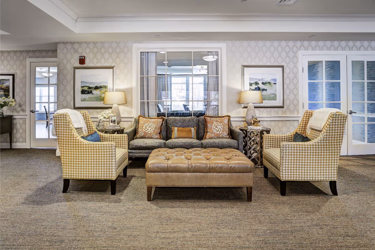 A cozy living room area with a gray sofa adorned with decorative pillows, two patterned armchairs with small cushions, and a large tufted leather ottoman in the center. The room features patterned wallpaper, two side tables with lamps and decorative items, framed artwork on the walls, and glass-paneled doors and windows in the background.