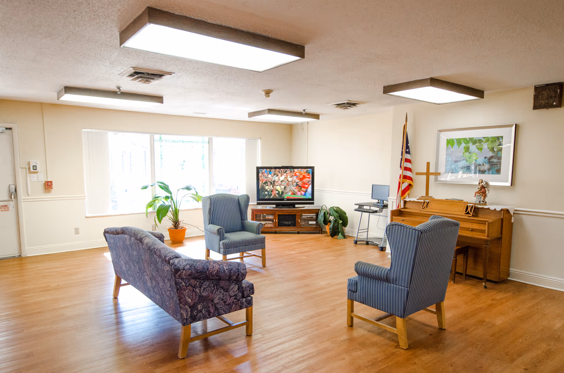 A bright common living area with wooden flooring featuring a floral-patterned sofa and two blue striped armchairs arranged around a television. There is a piano with a cross and a statue on top, an American flag, a computer on a stand, and some potted plants near a large window letting in natural light.
