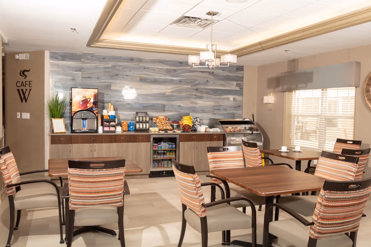 A cozy dining area with wooden tables and cushioned chairs featuring patterned backs. Against the wall, there is a counter with a coffee machine, snacks, and a small refrigerator stocked with beverages. The wall behind the counter has a modern wood panel design, and a sign reads 'Cafe W'. Large windows with blinds allow natural light to brighten the room.