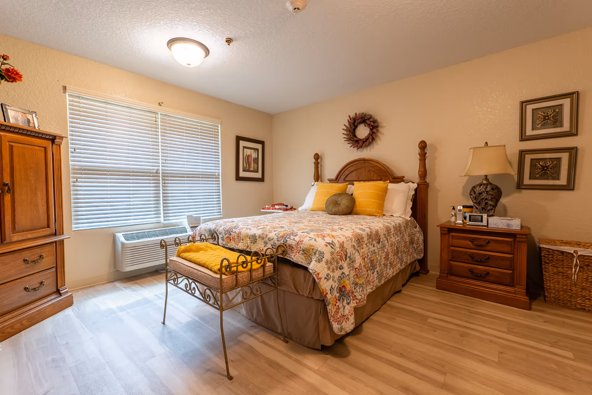 A cozy bedroom with a wooden bed frame and a floral quilt on the bed. The bed has three pillows, two yellow and one round green. There is a small bench with a yellow blanket at the foot of the bed. To the right of the bed is a wooden nightstand with a decorative lamp, a clock, and some personal items. On the wall above the nightstand are two framed pictures. To the left of the bed is a wooden dresser with drawers and a window with closed blinds letting in natural light. The floor is light-colored wood, and the walls are painted beige.