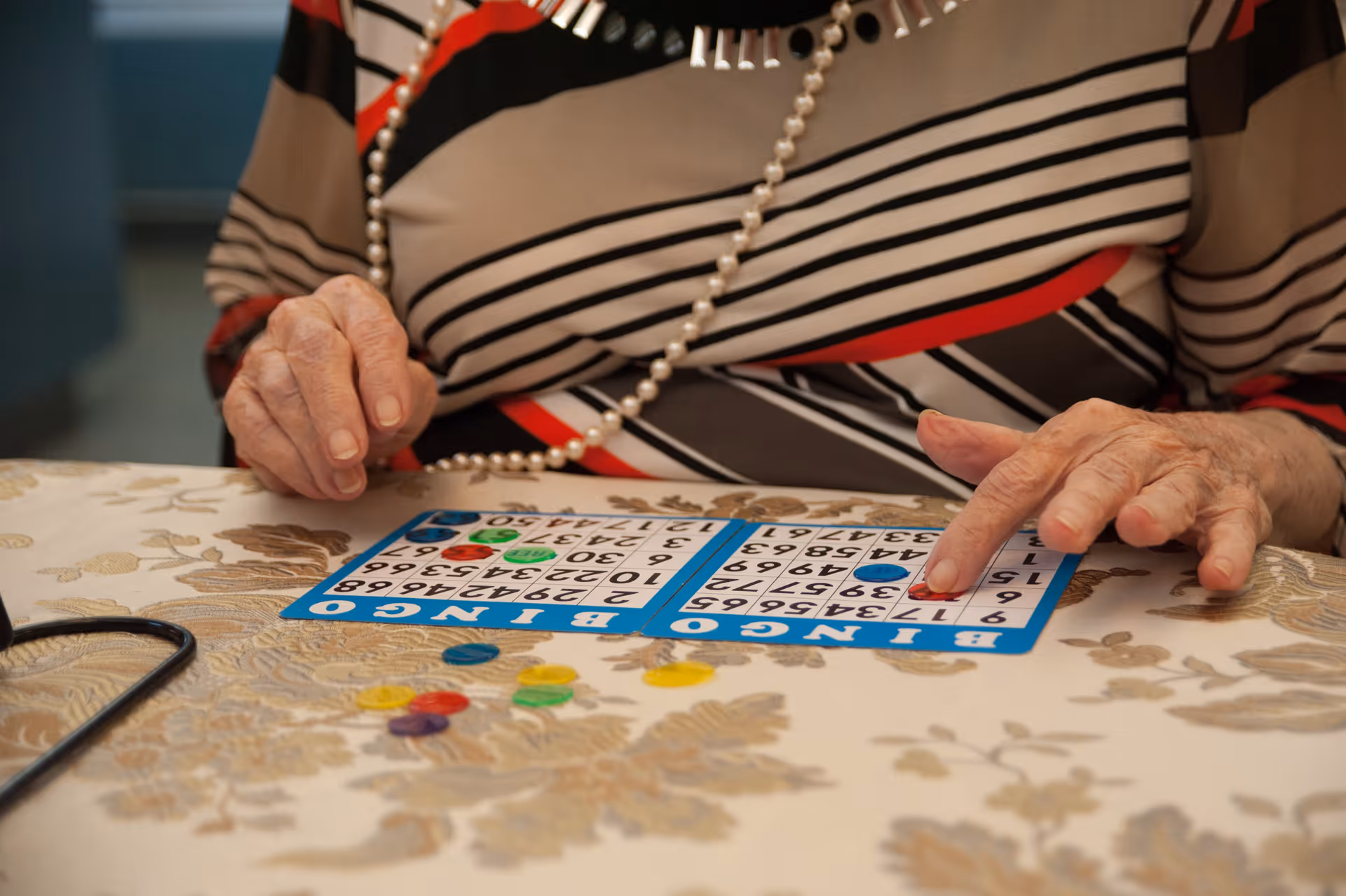 Close-up of an elderly person wearing a striped shirt and a pearl necklace, playing bingo with two bingo cards and colorful chips on a patterned tablecloth.