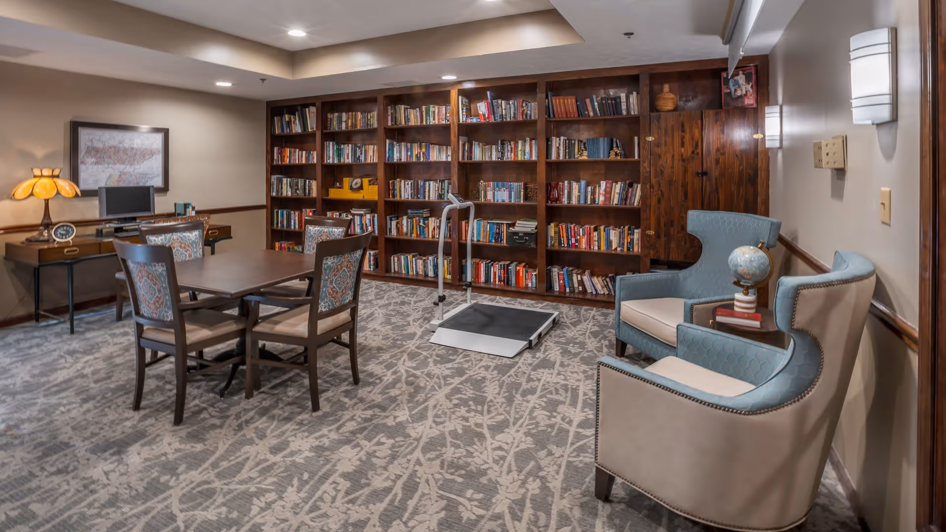 A cozy library room with a large wooden bookshelf filled with books, a table with four chairs in the center, two upholstered armchairs with a small side table holding a globe and books, a desk with a computer and a decorative lamp, and a patterned carpet covering the floor.