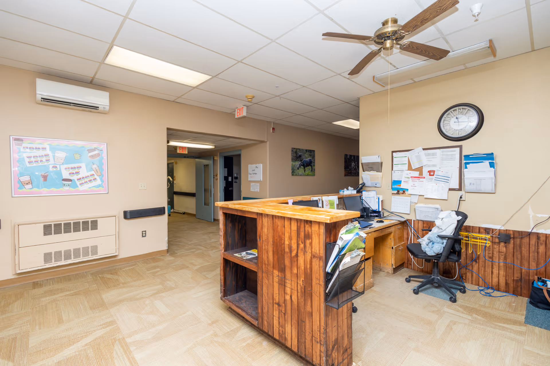 Reception area with a wooden desk, office chair, bulletin board, clock and ceiling fan in a facility hallway.