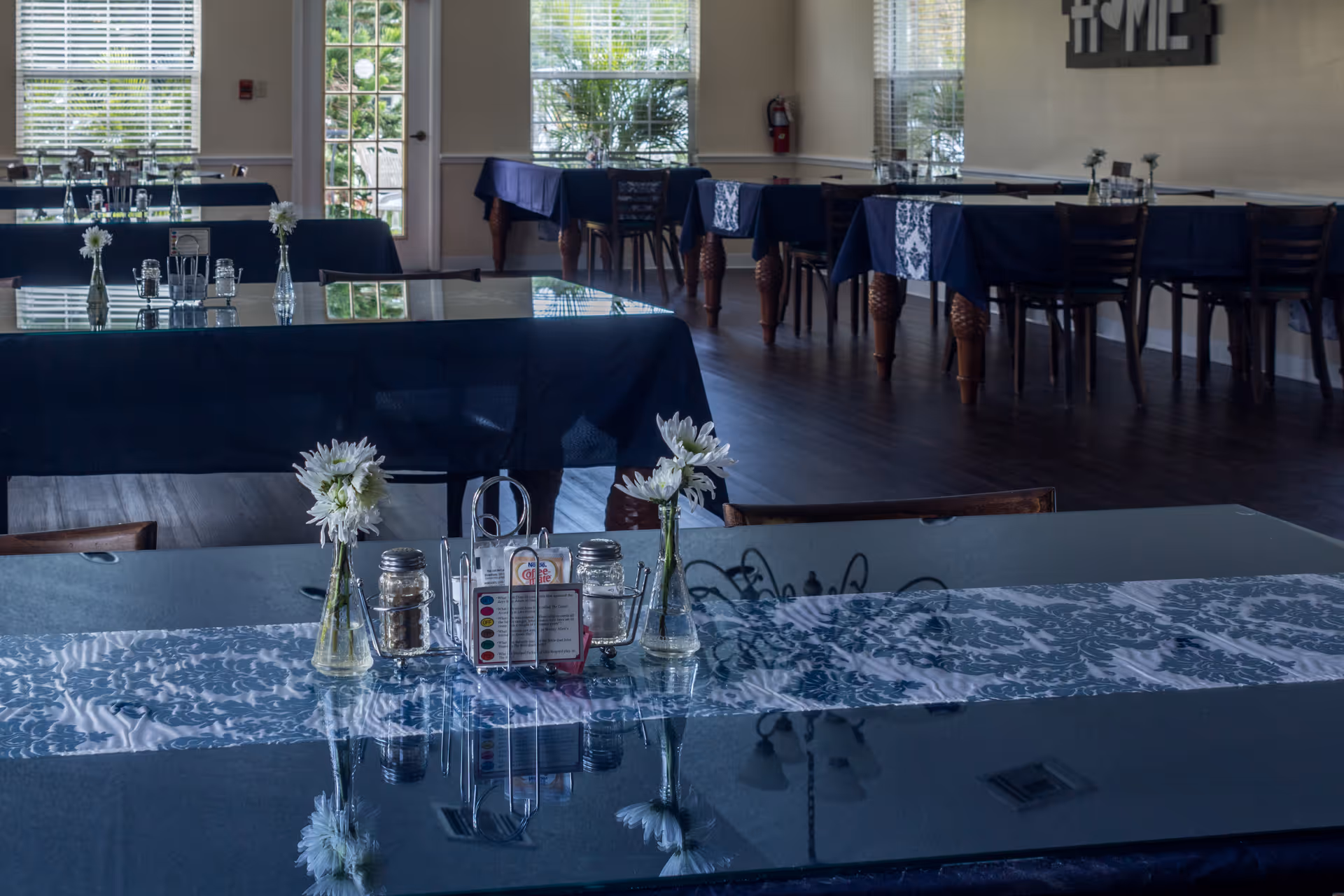 Interior view of a dining room with multiple tables covered in dark blue tablecloths and white patterned runners. Each table has small vases with white flowers and condiment holders. Large windows and a glass door allow natural light to brighten the room, which has dark wooden floors and chairs.