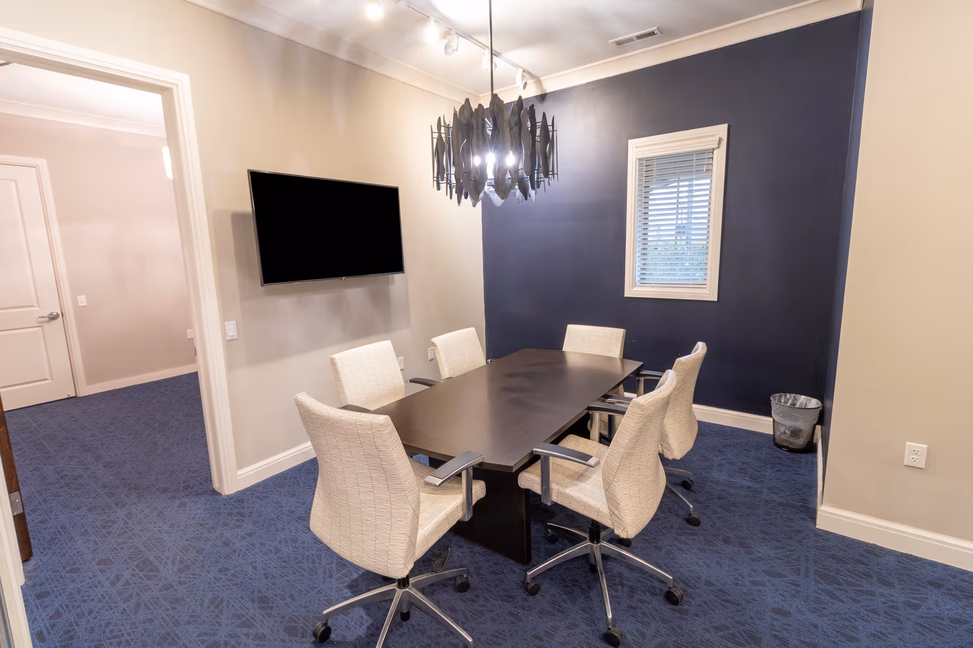 Small meeting room with a dark rectangular table, six wheeled upholstered chairs, a wall-mounted TV, and a navy accent wall with a window.