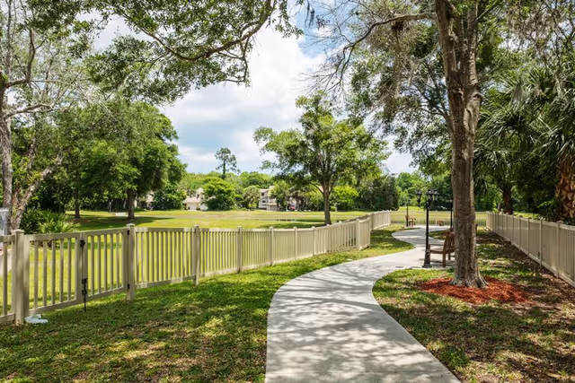 A winding concrete pathway bordered by a white fence on both sides, surrounded by green grass and trees under a partly cloudy sky in an outdoor garden area.