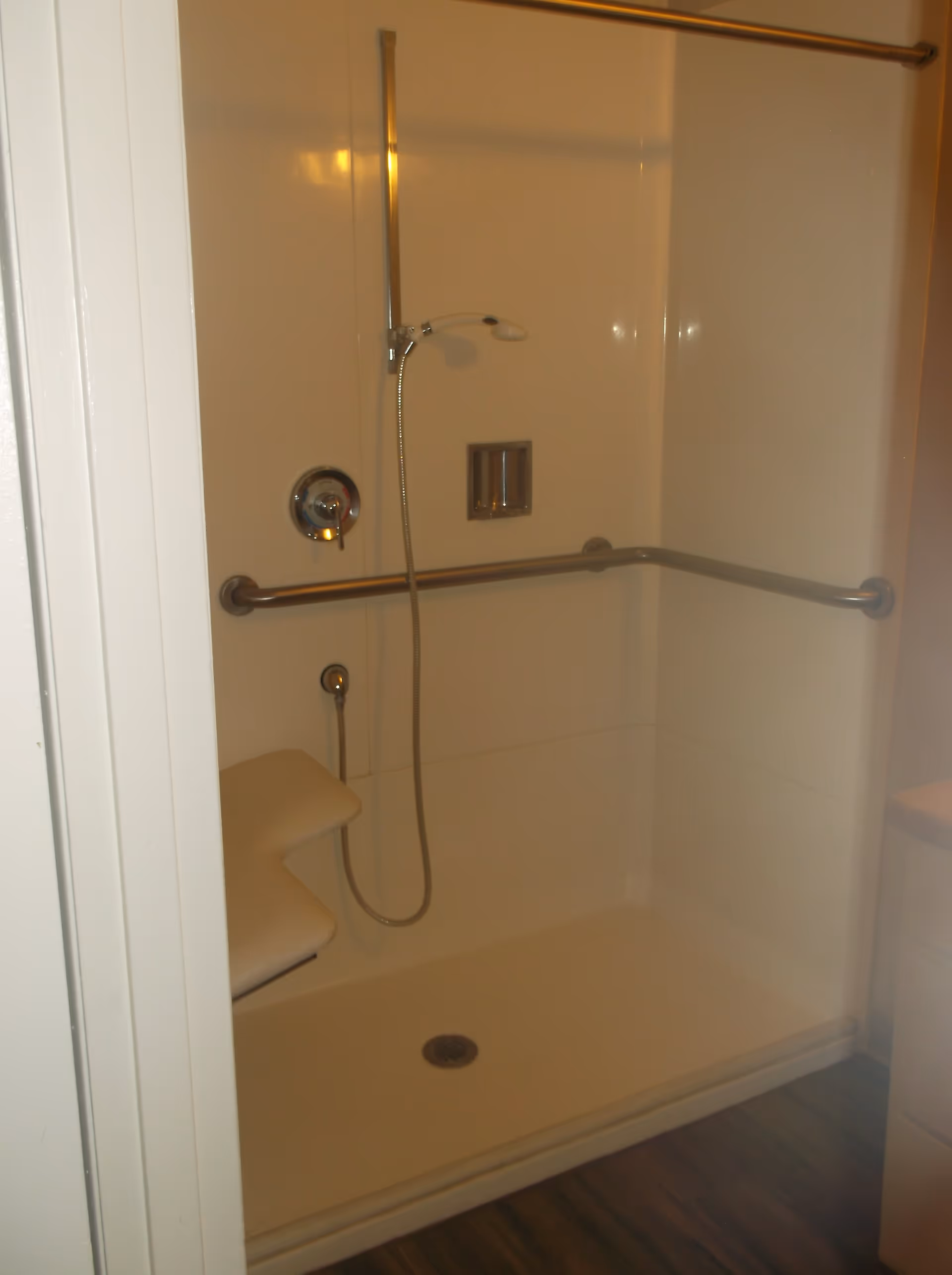 Accessible shower stall with a fold-down seat, handheld showerhead, and horizontal grab bars on two walls. The shower has a white interior and a wooden floor outside the shower area.
