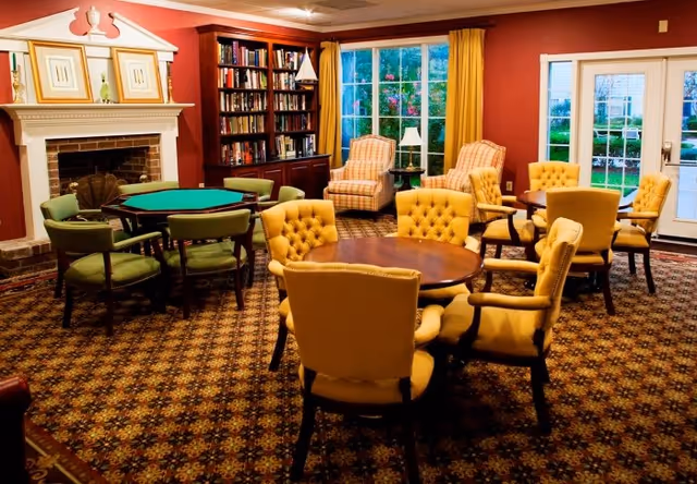 A cozy senior living common room with multiple round tables surrounded by upholstered chairs, a fireplace, bookshelves, and large windows.
