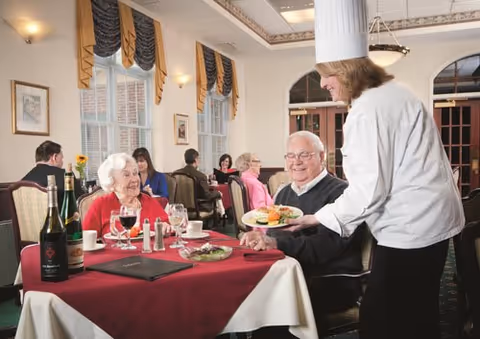 An elderly man and woman seated at a dining table in a restaurant setting with red tablecloths. A waitress wearing a white chef's hat and uniform is serving a plate of food to the man. Other diners are visible in the background, and the room has large windows with decorative curtains and framed pictures on the walls.