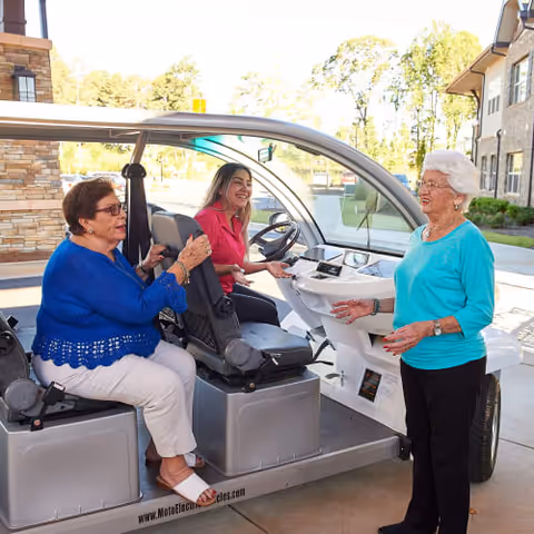 Three elderly women interacting near a small electric vehicle outside a senior living facility. One woman is standing and talking, while two others are seated inside the vehicle, smiling and engaged in conversation. The background shows part of the building and trees.