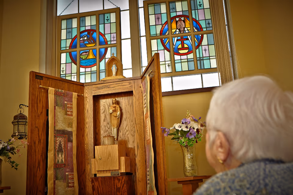 An elderly person in a chapel looks toward a wooden altar with a statue and stained-glass windows.