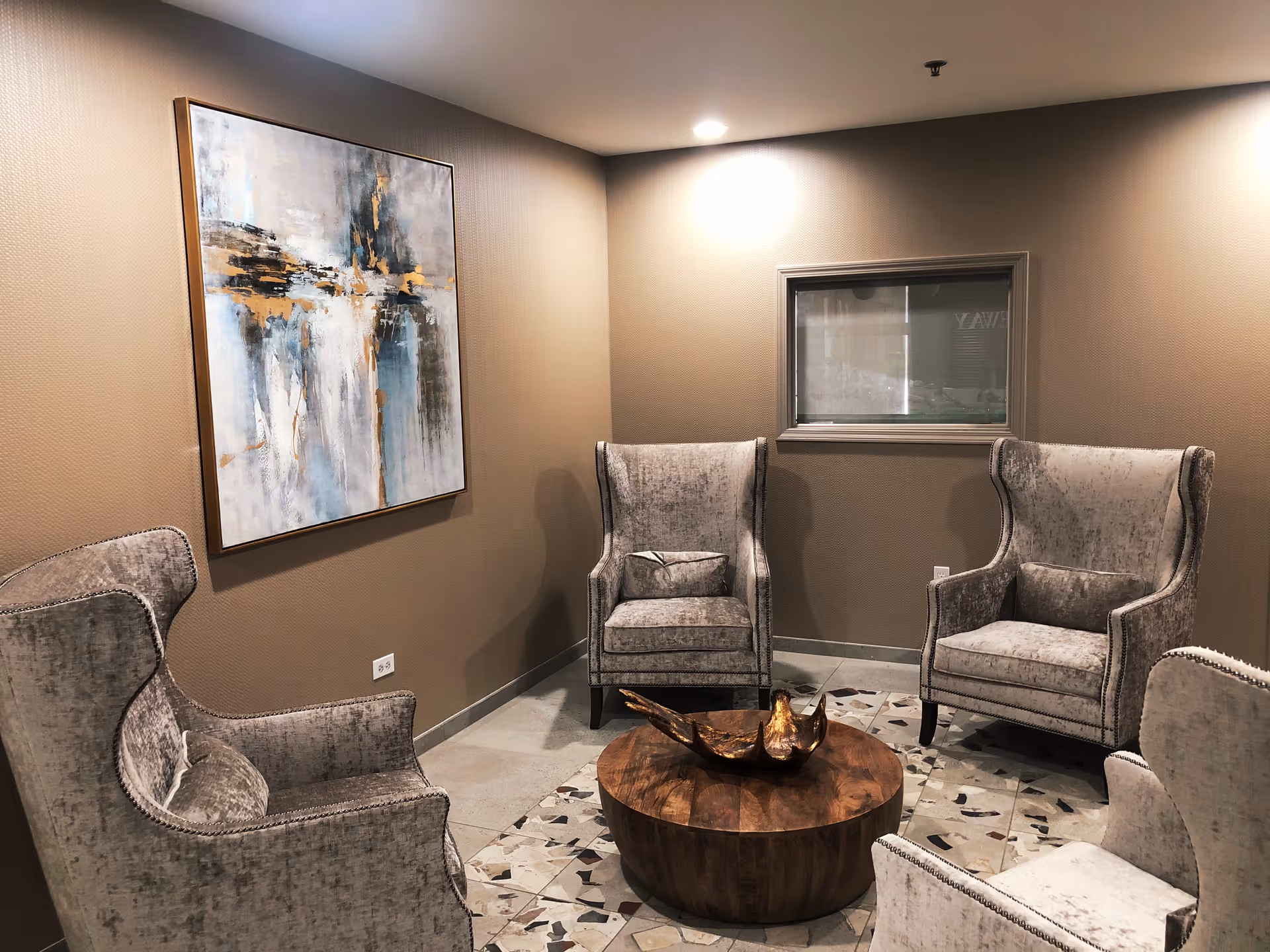 A small seating area with four gray upholstered armchairs arranged around a round wooden coffee table. The floor has a patterned tile design, and the walls are painted beige. A large abstract painting with blue, white, and gold tones hangs on one wall, and there is a small window on another wall.
