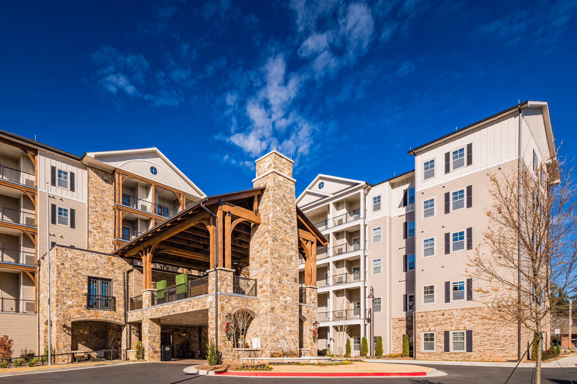 Exterior view of a multi-story senior living facility with stone and beige siding, featuring a large covered entrance with wooden beams and a stone chimney under a bright blue sky with scattered clouds.