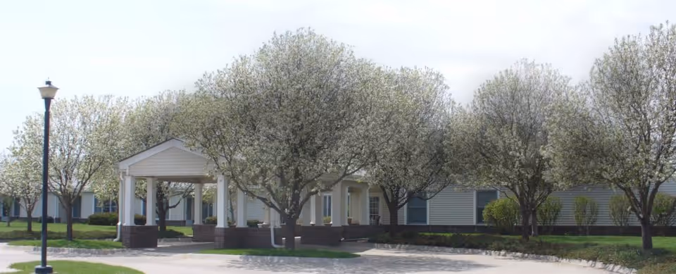 Exterior view of Prairie Village Retirement Center showing a single-story building with white siding, a covered entrance supported by white columns, and several trees with white blossoms lining the driveway.