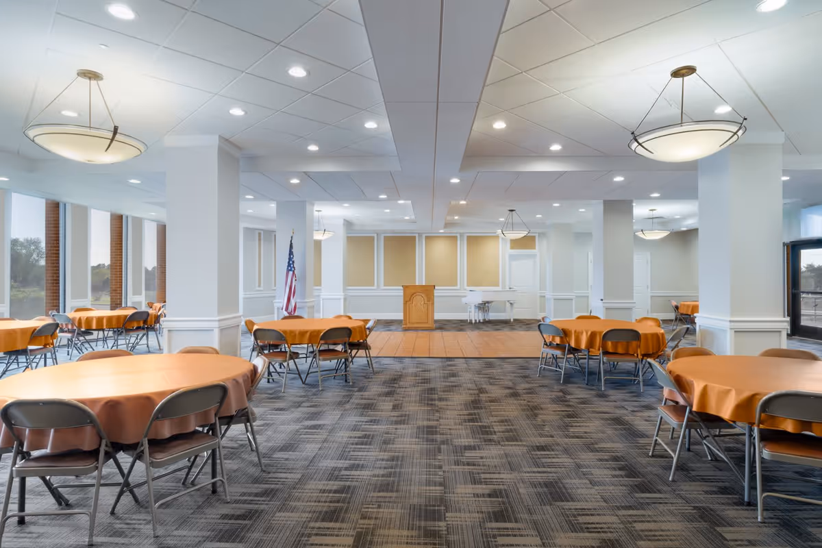 Spacious banquet-style dining room with round tables covered in orange tablecloths, folding chairs, a podium and an American flag.