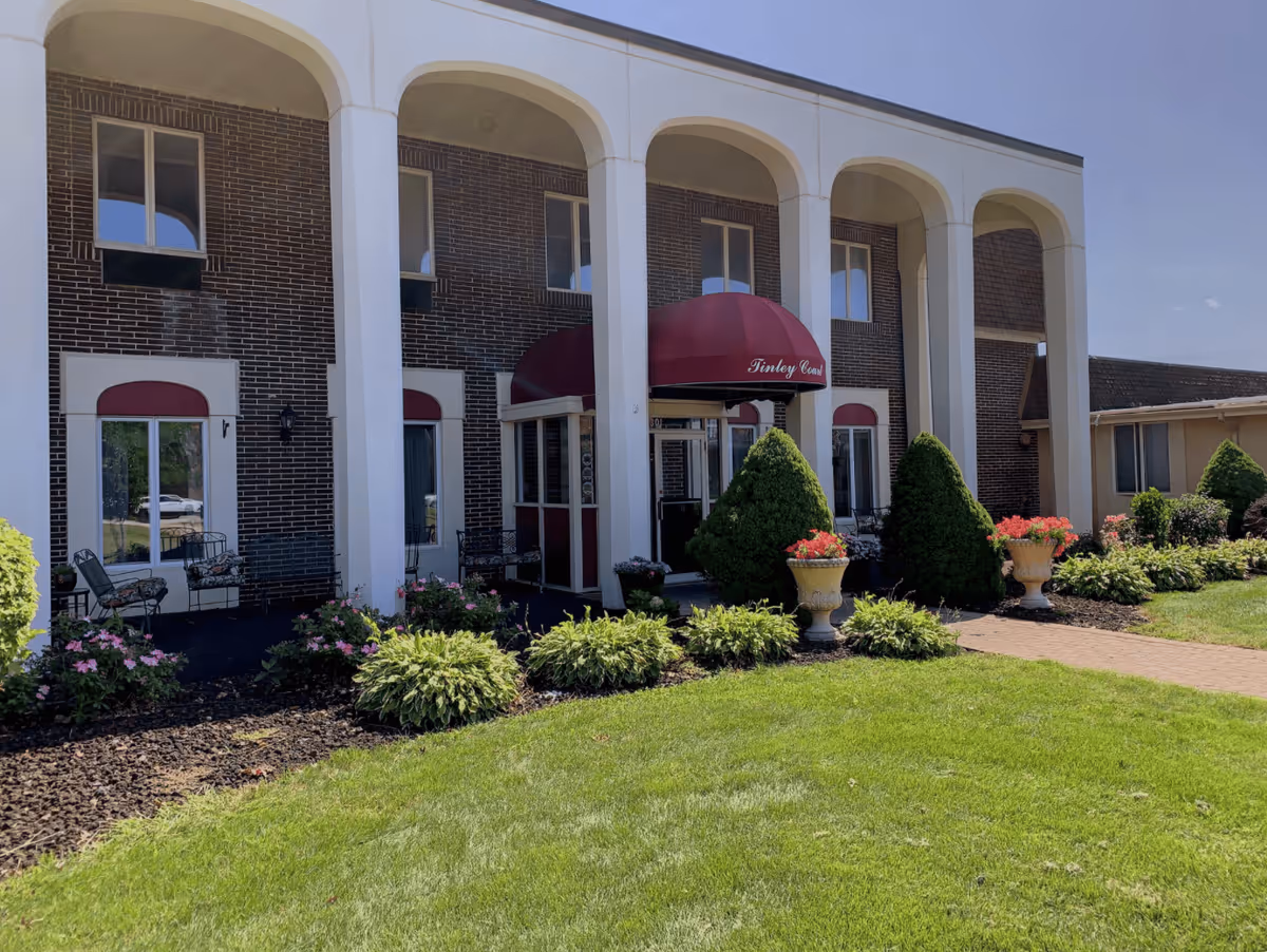 Exterior view of Tinley Court Catered Senior Living building with a brick facade and white columns. There is a red awning above the entrance with the name 'Tinley Court' on it. The entrance is surrounded by well-maintained landscaping including green bushes, flowering plants, and a neatly trimmed lawn.