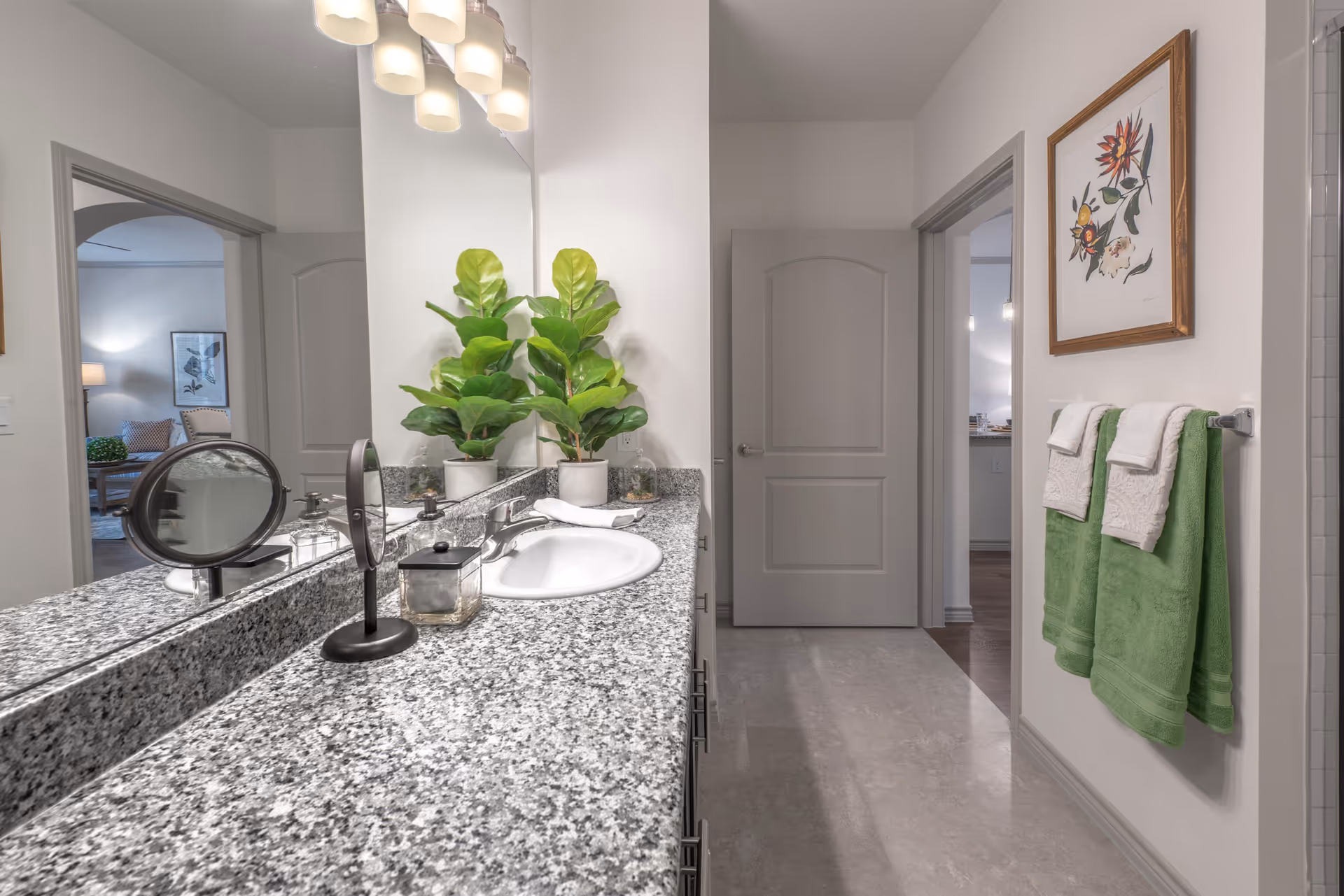 A bathroom with a granite countertop featuring a sink, a small round mirror, soap dispenser, and a potted plant. Above the sink is a large mirror with three light fixtures. On the right wall, two green towels with white decorative towels hang on a towel rack beneath a framed floral artwork. The bathroom floor is light-colored, and there are two doorways visible, one leading to a living room area and the other to another room.