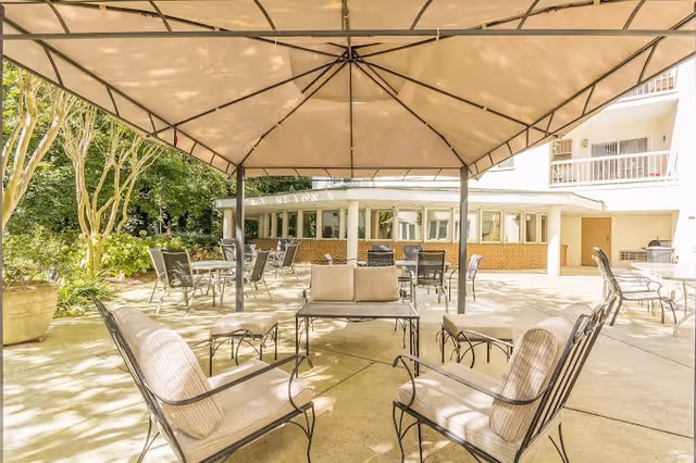 Outdoor shaded patio with cushioned metal chairs and tables under a large canopy beside a residential building.
