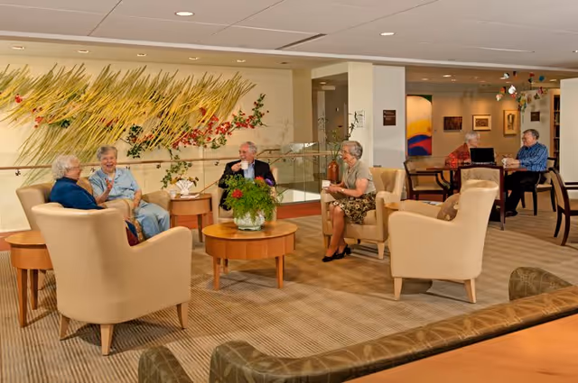 A group of elderly people sitting and chatting in a spacious, well-lit common area with beige armchairs and wooden tables. The room features a decorative wall installation with plants and flowers, carpeted floors, and additional seating with two people conversing at a table in the background.