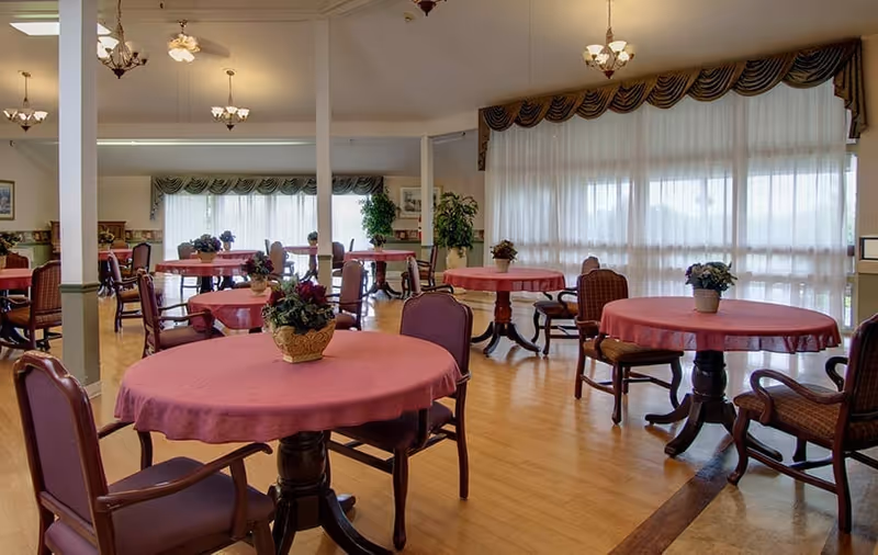 Dining room with round tables covered in pink tablecloths, chairs, potted centerpieces, and large windows with sheer curtains.