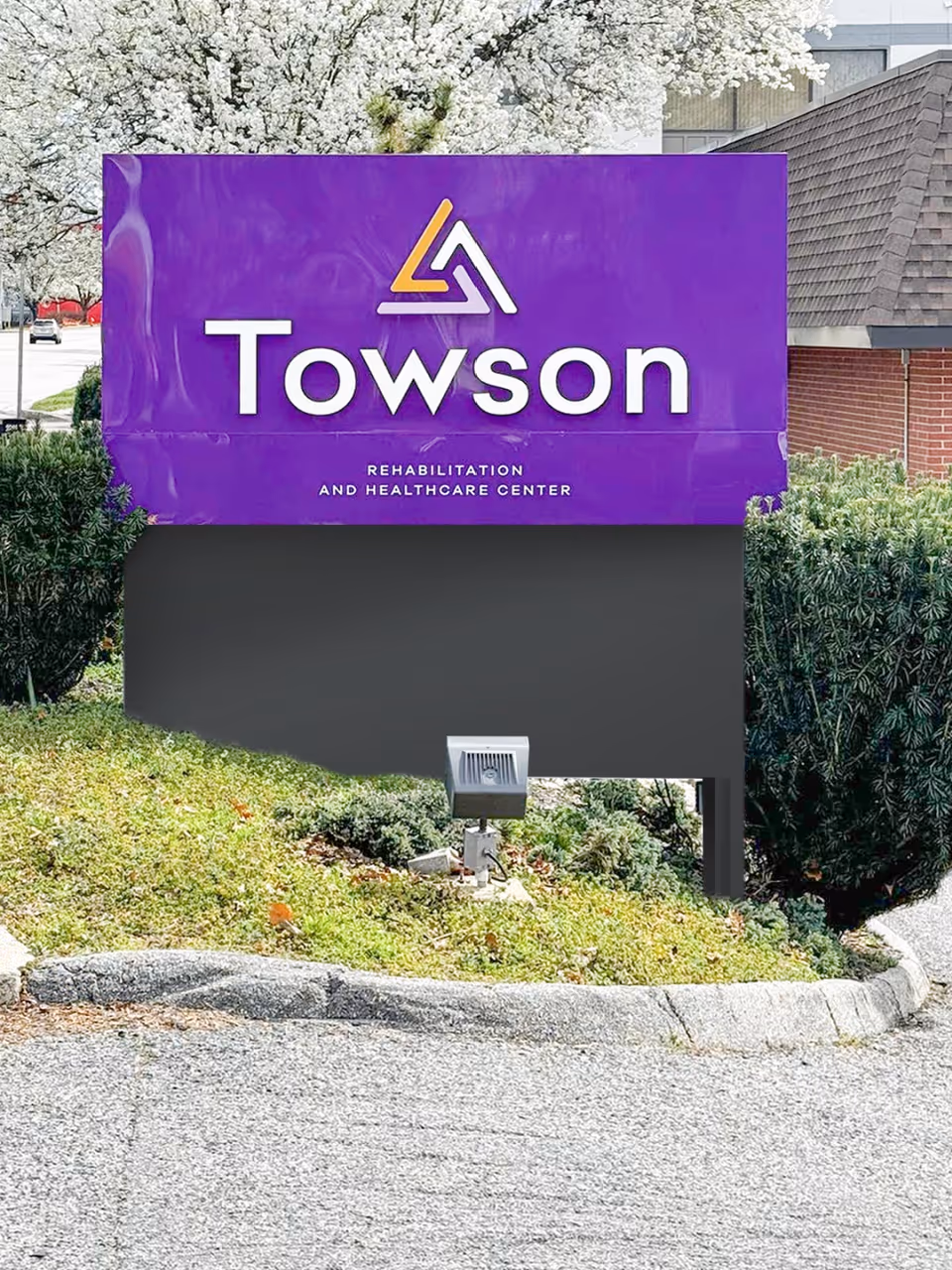 Outdoor sign for Towson Rehabilitation and Healthcare Center with a purple background and white text, surrounded by greenery and a tree with white blossoms in the background.