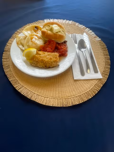 A white plate on a woven placemat with a serving of fried fish with a lemon wedge, a bread roll with butter, cooked tomatoes, and pierogies. Next to the plate is a folded white napkin with a fork, knife, and spoon on a dark blue tablecloth.