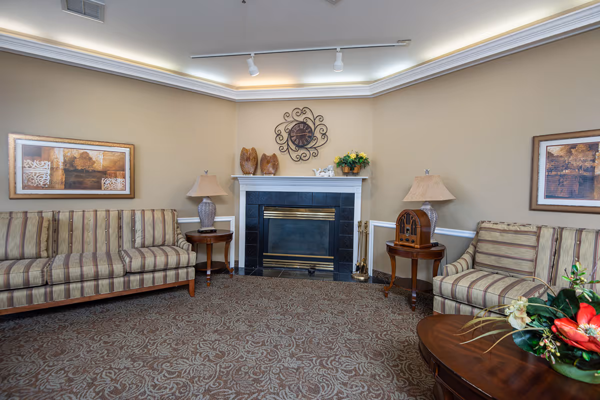A cozy living room area with two striped sofas facing each other, a fireplace with decorative items on the mantel, two side tables each with a lamp, framed artwork on the walls, and a patterned carpet. There is also a round wooden coffee table with a floral arrangement in the foreground.