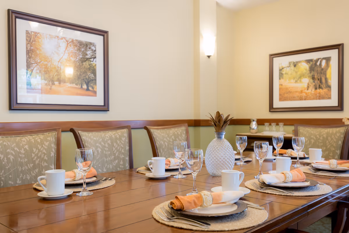 A neatly set dining table with plates, napkins, glassware and a decorative vase in a well-lit dining room with framed artwork on the walls.