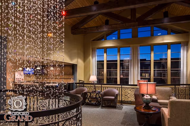 Comfortable assisted-living lounge with upholstered chairs and sofas, table lamps, large windows, decorative railing and a hanging crystal light installation.