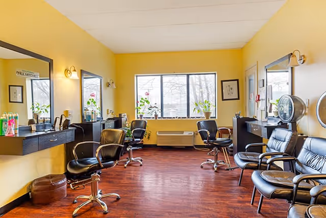 Bright salon-style room with multiple black styling chairs, mirrors, and a large center window overlooking trees.