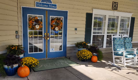 Entrance of Sumter Grove Senior Living with double blue doors decorated with autumn wreaths, a 'Welcome Home' sign above the doors, potted plants, pumpkins, and two white rocking chairs with blue patterned cushions on the porch.
