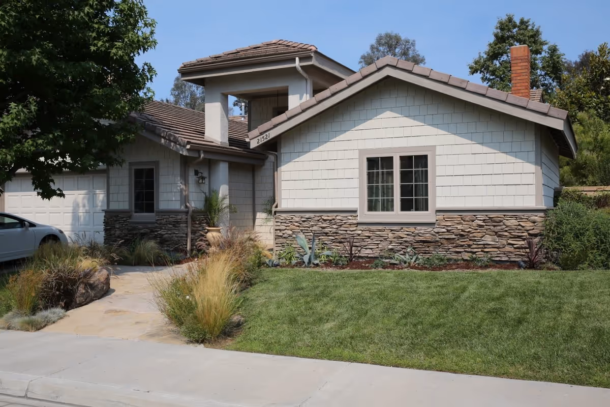 Front exterior view of a single-story house with a stone and shingle facade, a tiled roof, a green lawn, and landscaped plants along the walkway leading to the entrance. A white car is partially visible parked on the left side.