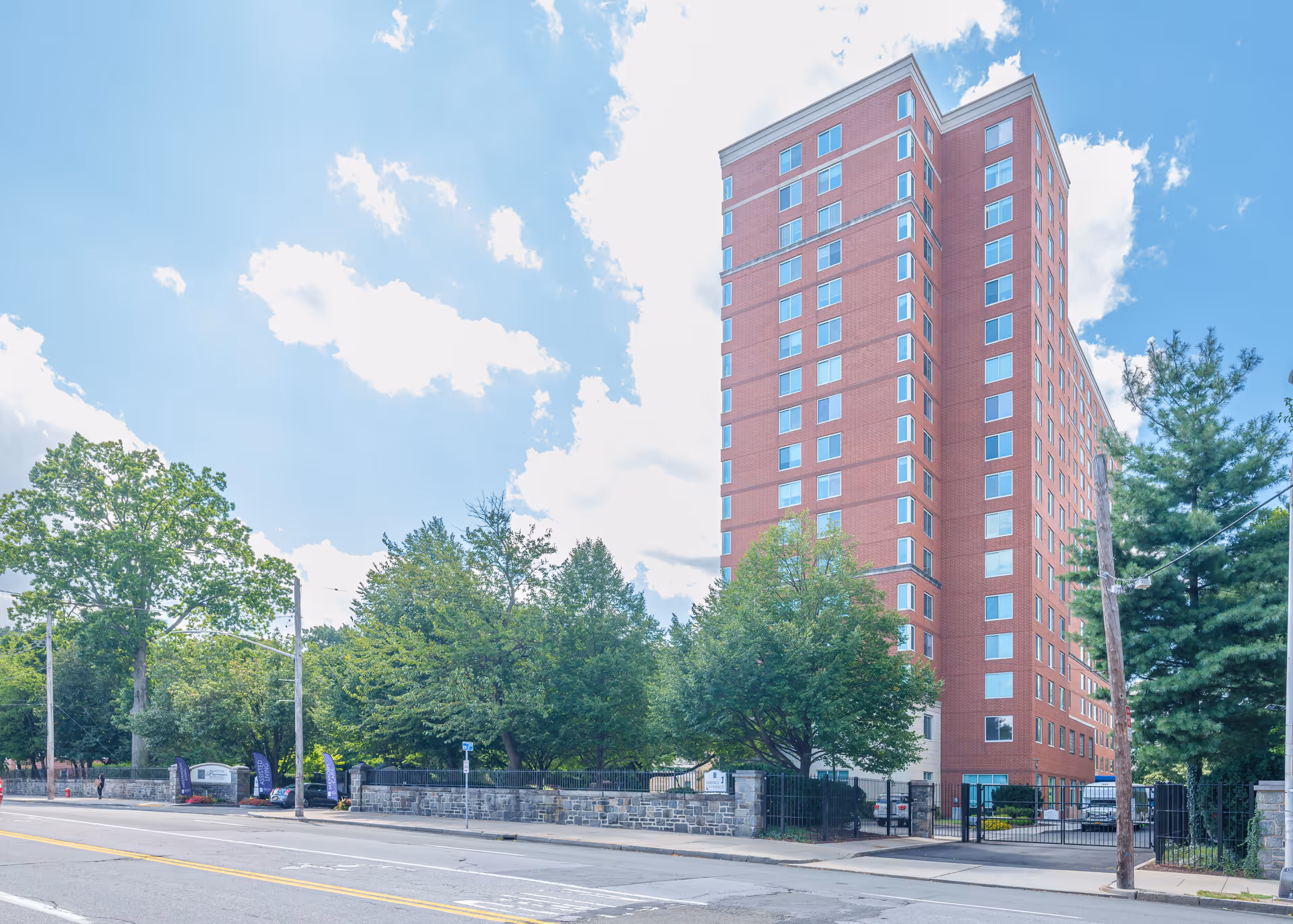 Tall multi-story red brick building with many windows, surrounded by trees and a stone fence, under a partly cloudy blue sky.