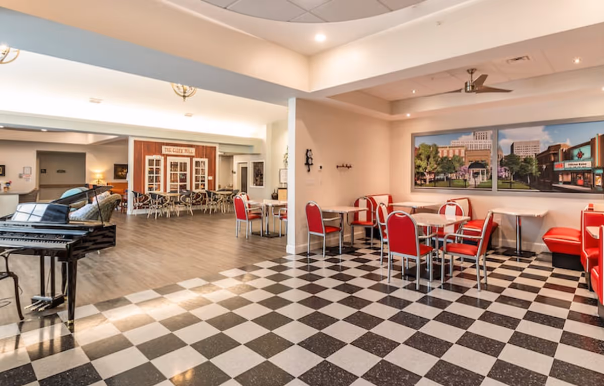 Interior view of a senior living facility common area featuring a black and white checkered floor section with red chairs and tables, a mural of a cityscape on the wall, and an adjacent open space with wooden flooring, additional seating, and a black grand piano.