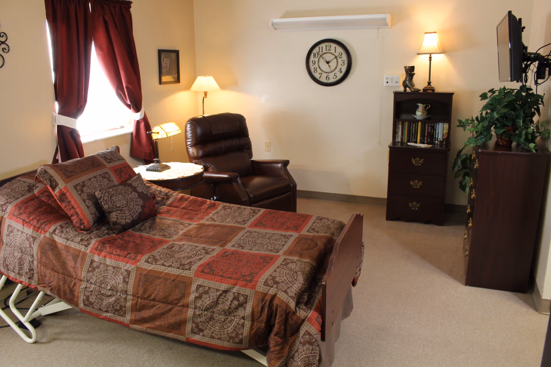 A cozy bedroom with a single bed covered in a patterned brown and red quilt, a brown leather recliner chair next to a small round table with a lamp, a wall clock above a small bookshelf with books and decorative items, a flat-screen TV mounted on the wall, and a potted plant on a wooden dresser. The room has beige walls and carpet, and a window with red curtains.