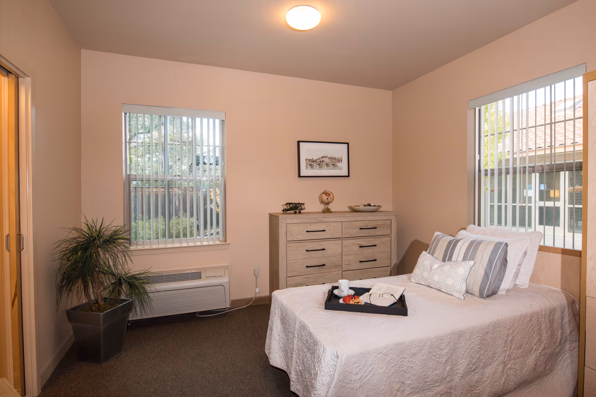 A cozy bedroom in Meadow Oaks of Roseville featuring a neatly made bed with white bedding and decorative pillows. A black tray with a cup, an open book, and some fruit rests on the bed. The room has two windows with vertical blinds, a wooden dresser with decorative items on top, a potted plant in the corner, and soft beige walls.