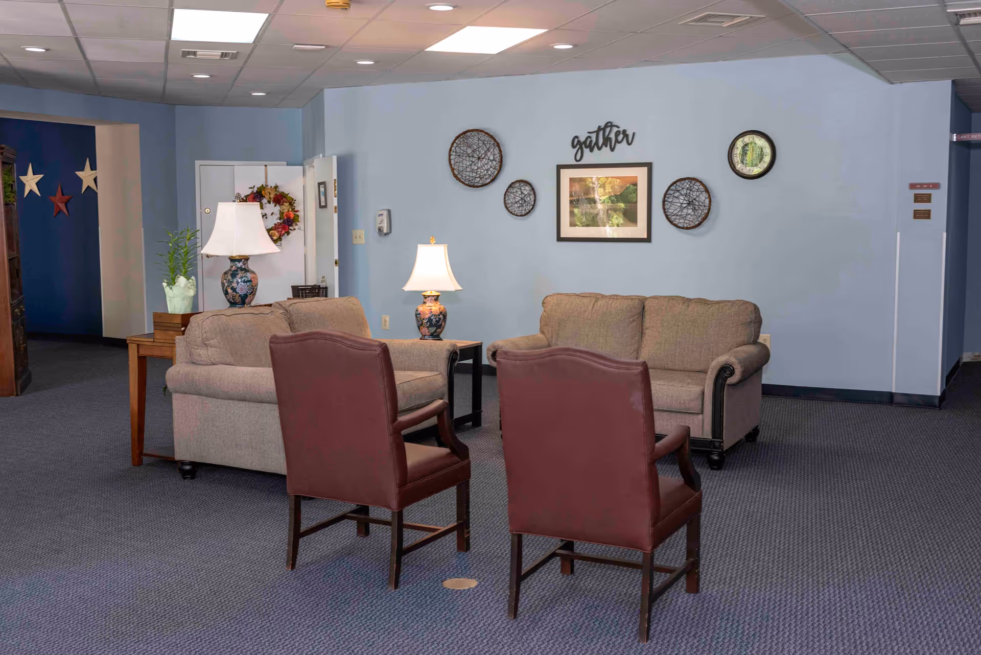 A cozy seating area in a senior living facility with two beige sofas, two maroon armchairs, two table lamps with floral bases, and wall decorations including a framed picture, circular woven art pieces, and the word 'gather' mounted on a light blue wall.